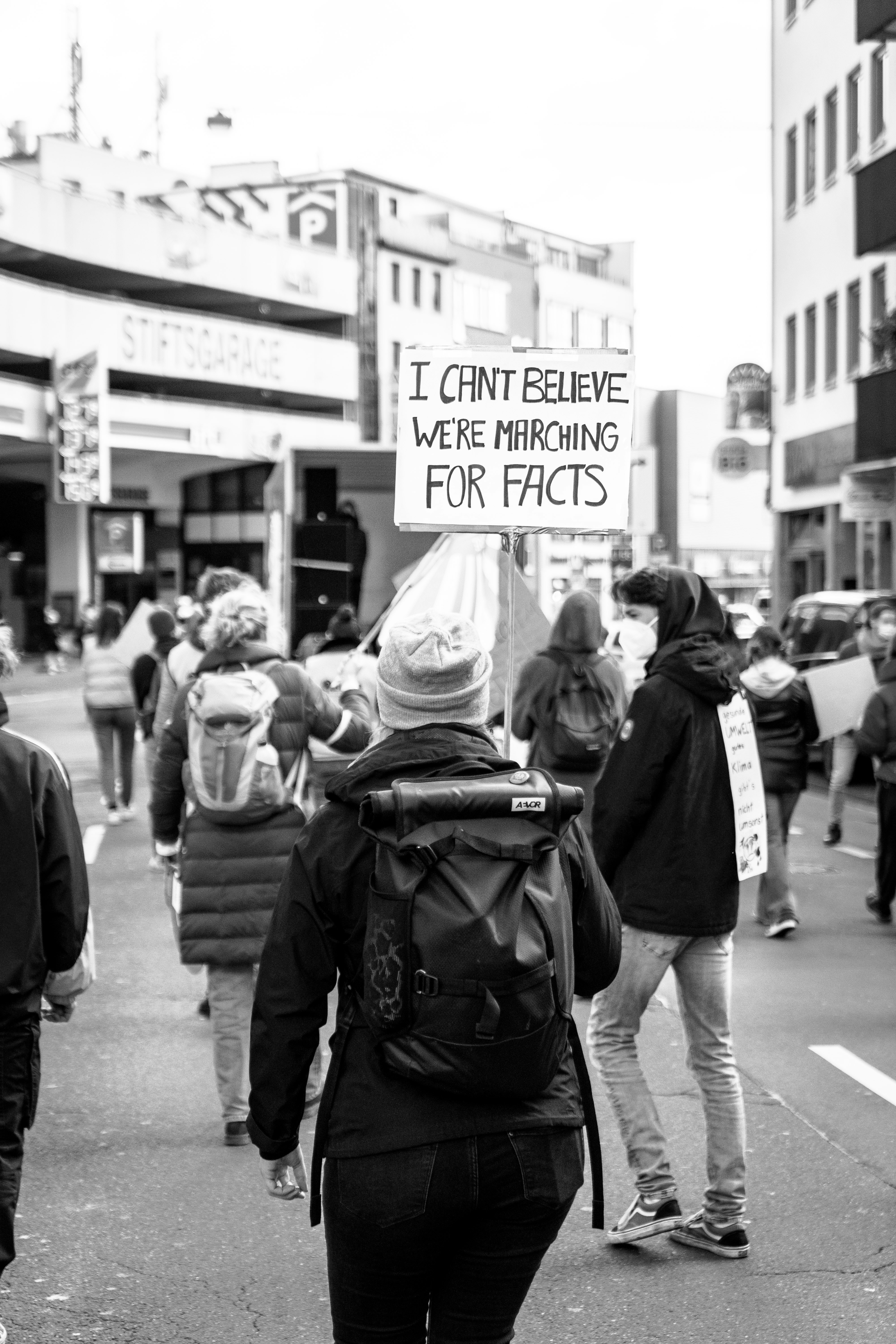 Black-and-white photo of protesters marching away from the camera, with a sign reading "I CAN'T BELIEVE WE'RE MARCHING FOR FACTS"